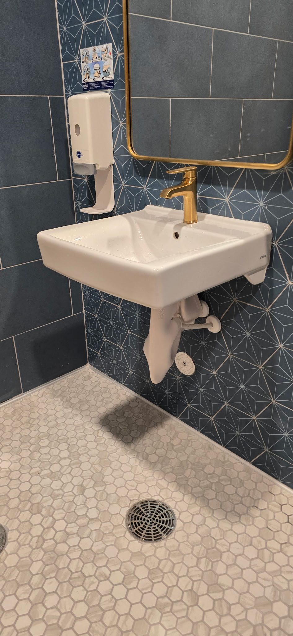 Wall-mounted white lavatory sink with a brass faucet installed against blue tile in a finished commercial restroom