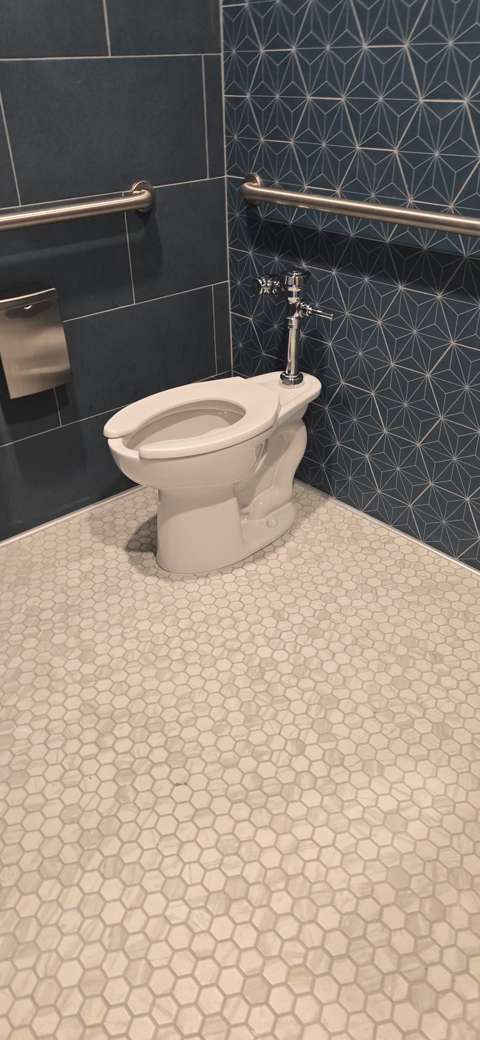 ADA-height commercial toilet installed beside stainless steel grab bars on hexagon tile flooring