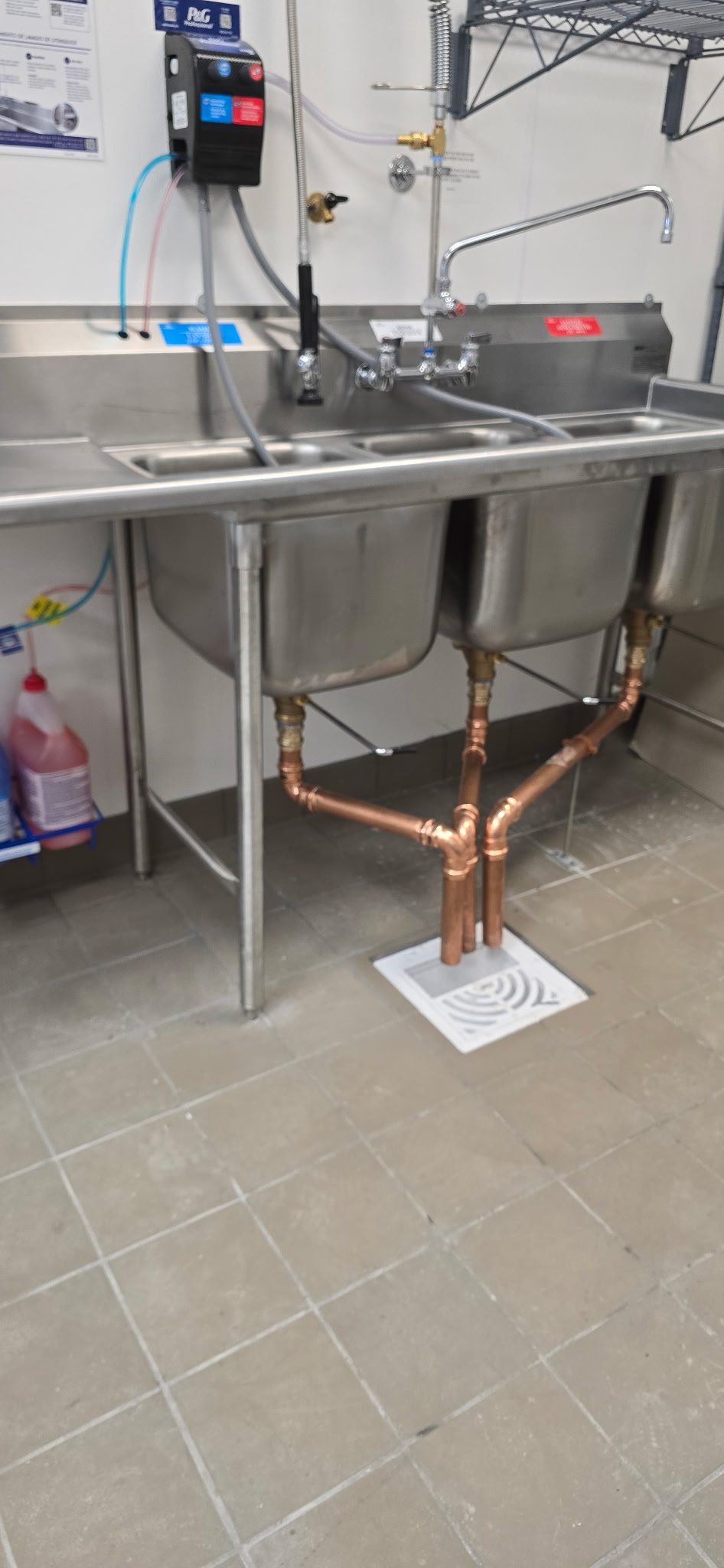 Copper waste piping from a three-compartment sink discharging into a white indirect floor sink drain