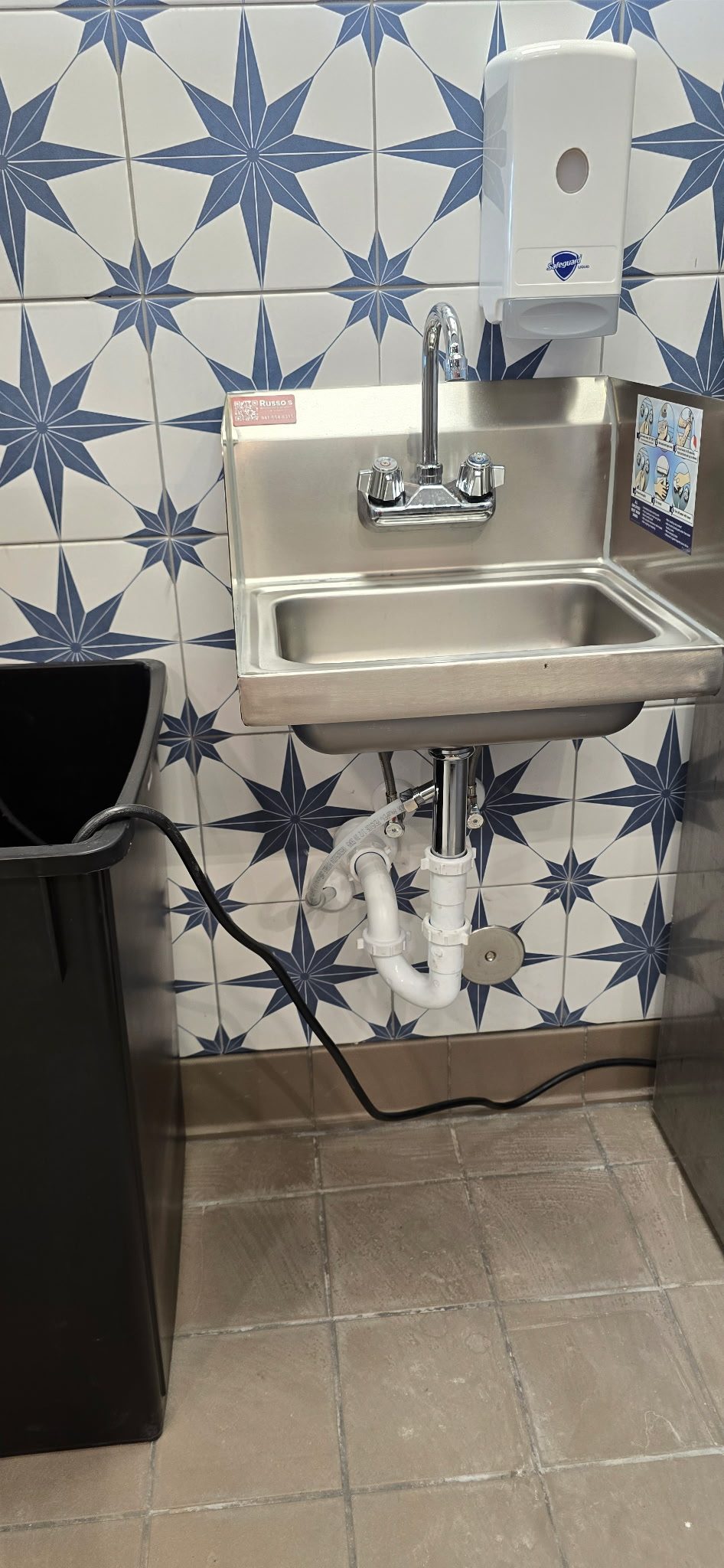 Stainless steel wall-mounted hand sink with new P-trap and supply lines against patterned blue tile